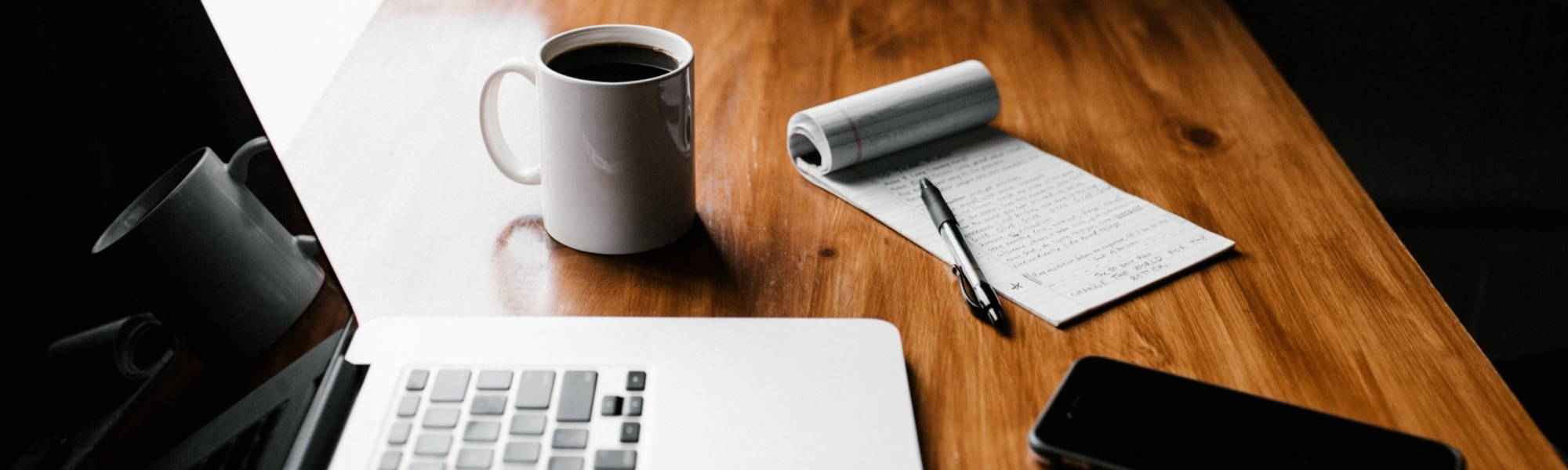 A lap top sits open on a desk alongside a mug and notebook and pen