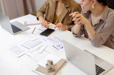 Two women collaborate at a table, surrounded by papers and laptops, engaged in discussion and work.