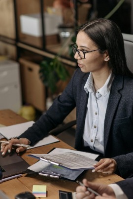 A woman in glasses and a gray jacket sits confidently at a table,  holding note pad