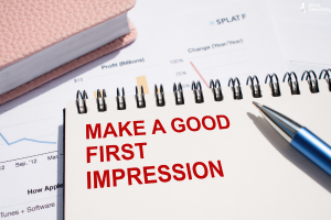 A white notepad with the writing 'make a good first impression' on it and a pen on a desk with other paper and books.