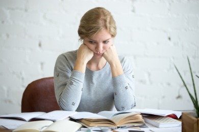 A woman appears frustrated at work, seated at a table filled with books and papers, reflecting her stress.