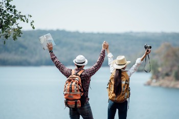 Two hikers with backpacks stand on a cliff, gazing at a serene lake surrounded by lush greenery.