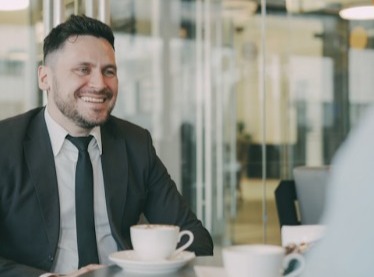 A person in a business suit sitting at a table in a modern café or office setting, with coffee cups in front of them during a conversation.
