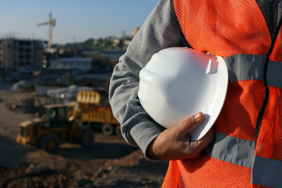 A man in an orange vest and hard hat holds a white hard hat, ready for construction work.
