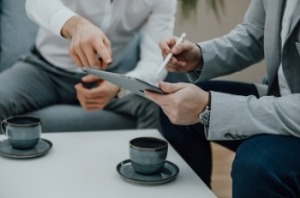 Two men in business attire sit on a couch, discussing cover letter writing while using a tablet.