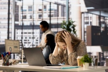 A woman at a desk, visibly stressed, with her head in her hands, reflecting workplace stress.