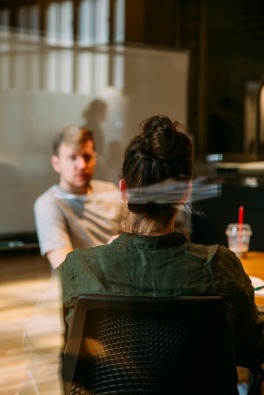 Two people are engaged in a conversation in an office, seen through a reflective glass. The setting is casual, with a relaxed and focused atmosphere.