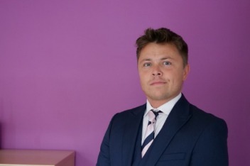 Man in a dark suit and striped tie stands against a vibrant purple background, giving a confident and professional appearance.