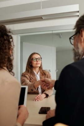 A woman in glasses and a blazer shakes hands with a man across a desk, while another person with a tablet observes, suggesting a successful meeting.