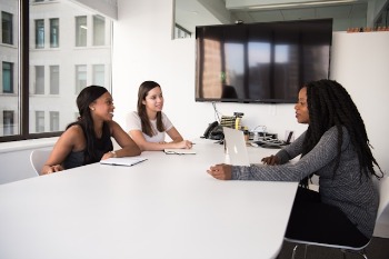 Three people sit around a white conference table in a modern meeting room; one works on a laptop while the others listen, with a large wall‑mounted screen behind them.