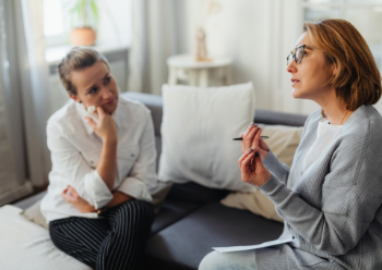 Two women engaged in conversation while sitting on a couch, sharing smiles and enjoying each other's company.