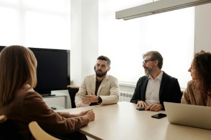 A group of four people sit around a conference table in a bright office. One person speaks while others listen attentively, creating a collaborative atmosphere.