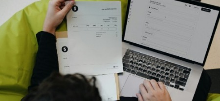 A person is sitting on a green beanbag, holding a document in one hand and typing on a laptop with the other. The scene conveys focused work.