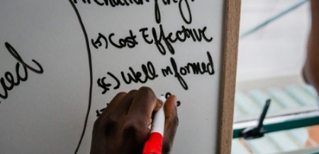 A hand writing on a whiteboard with a red marker. The board features phrases like "Cost Effective" and "Well Informed." The scene conveys focus and planning.