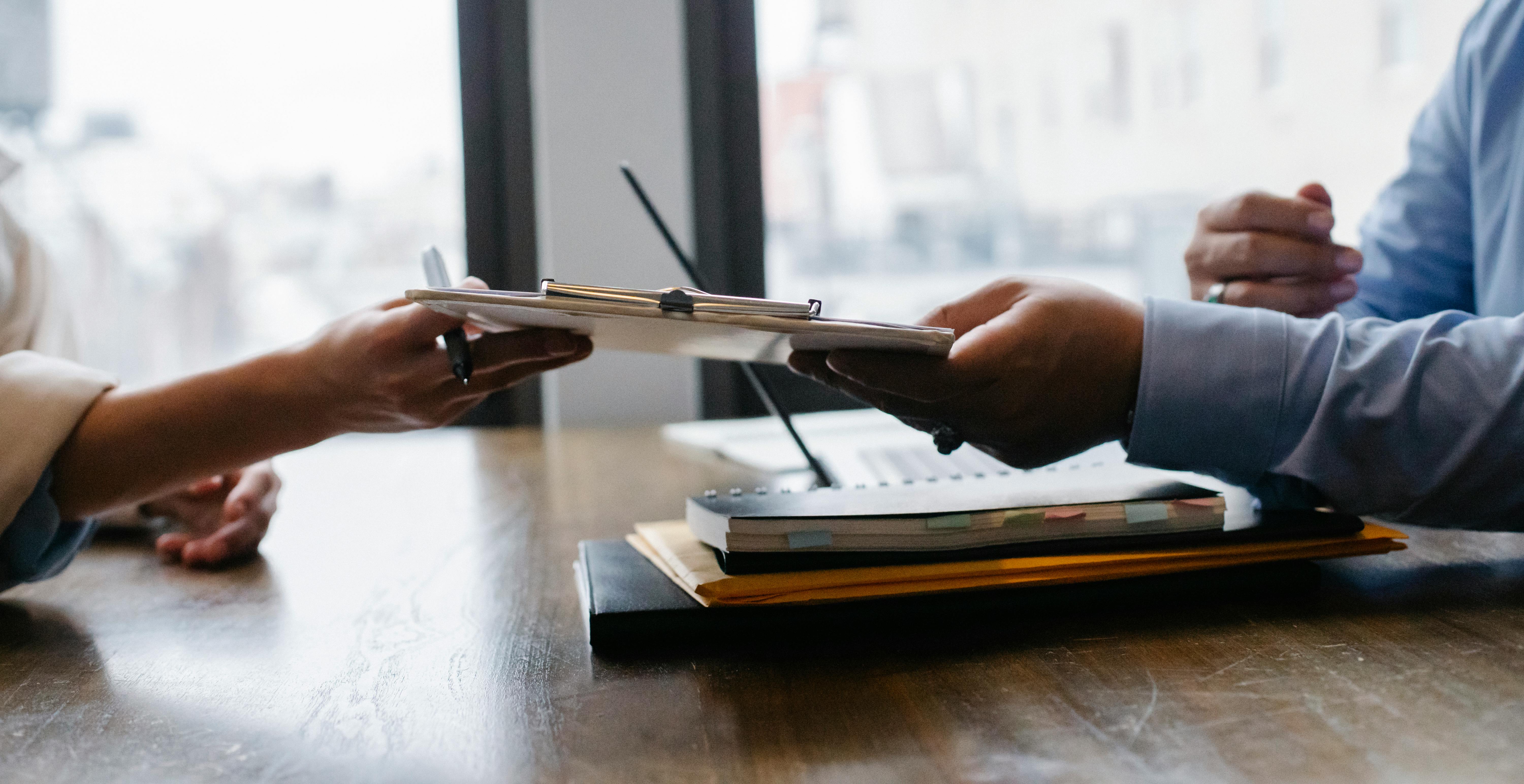 Two people exchange a clipboard across a table, suggesting teamwork or collaboration. Documents and a notebook are visible, creating a professional tone.