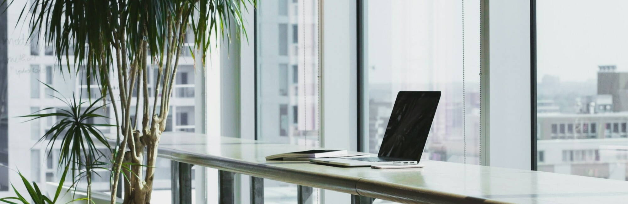A laptop sits on a table in front of a large window, allowing natural light to illuminate the workspace.