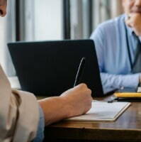 A man and woman sit at a desk; the man is interviewing for an accounting position using a laptop.